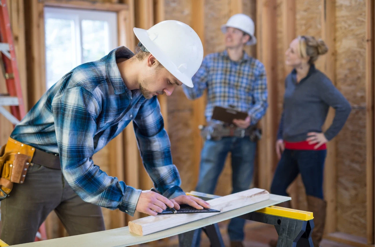 Construction worker in a white hard hat measuring wood at a renovation site, with colleagues discussing plans in the background.